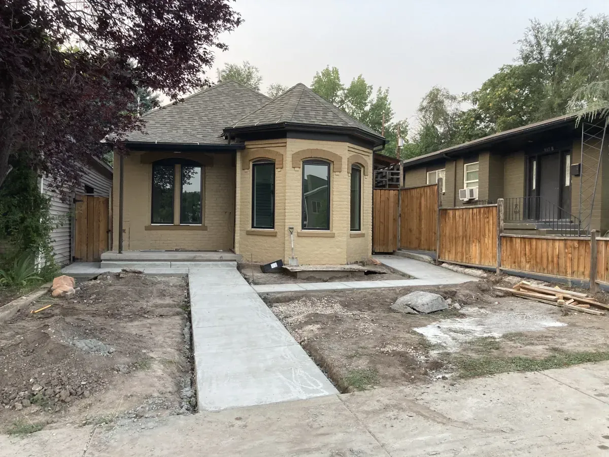 Small beige house with a front walkway and unfinished yard, flanked by neighboring homes.