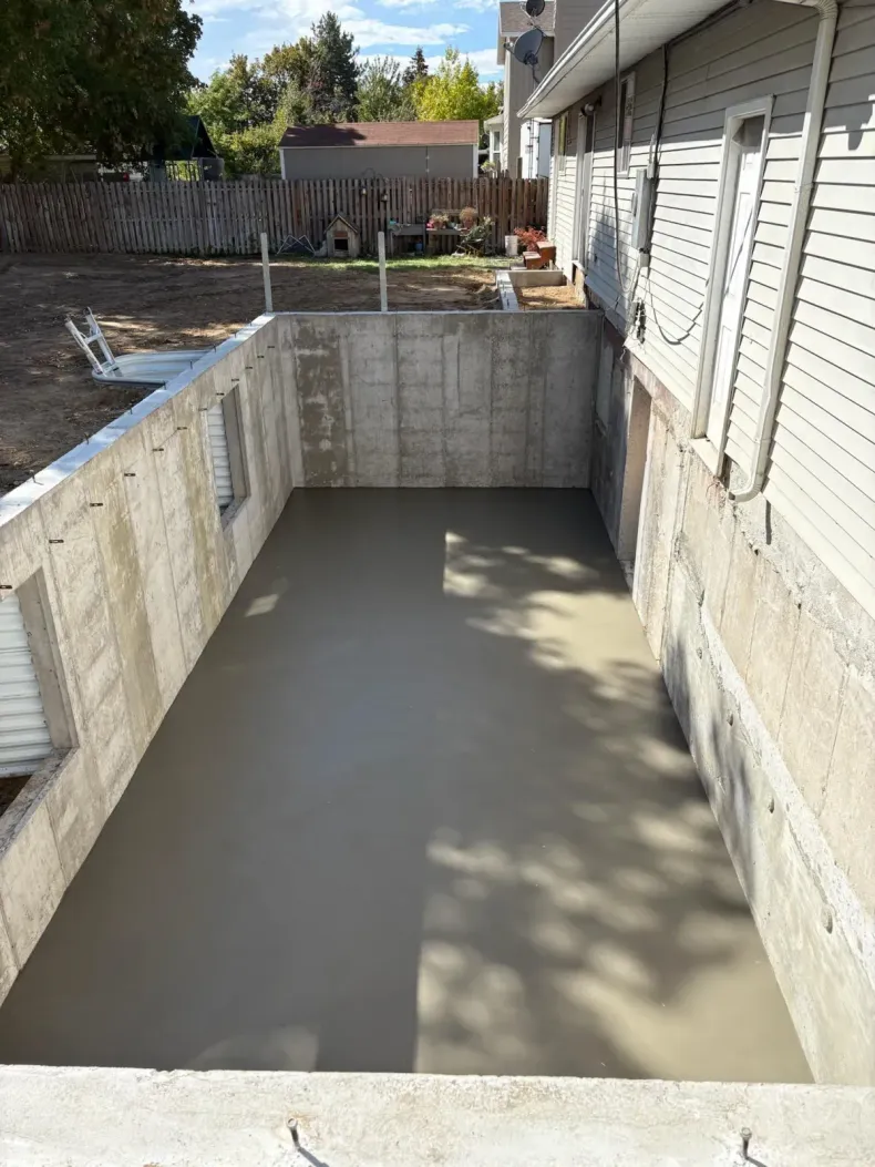 Rectangular concrete basement trench beside a house, with freshly poured smooth floor and retaining walls