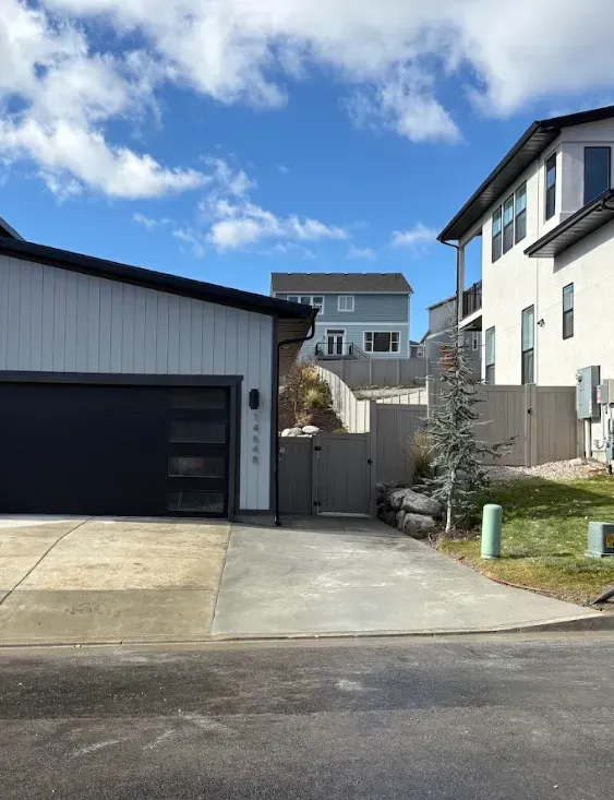 Suburban driveway between a white garage and a house, with a gate, tree, and blue sky.