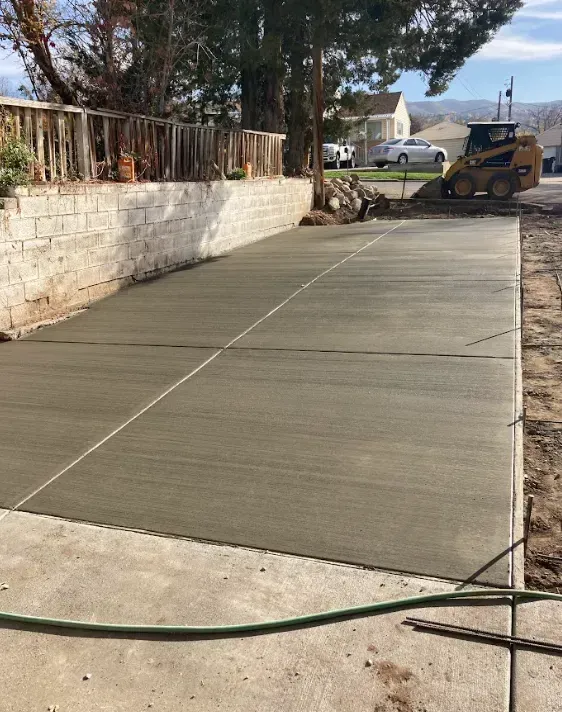 Freshly poured concrete driveway beside a stone wall, with a skid steer in the background.