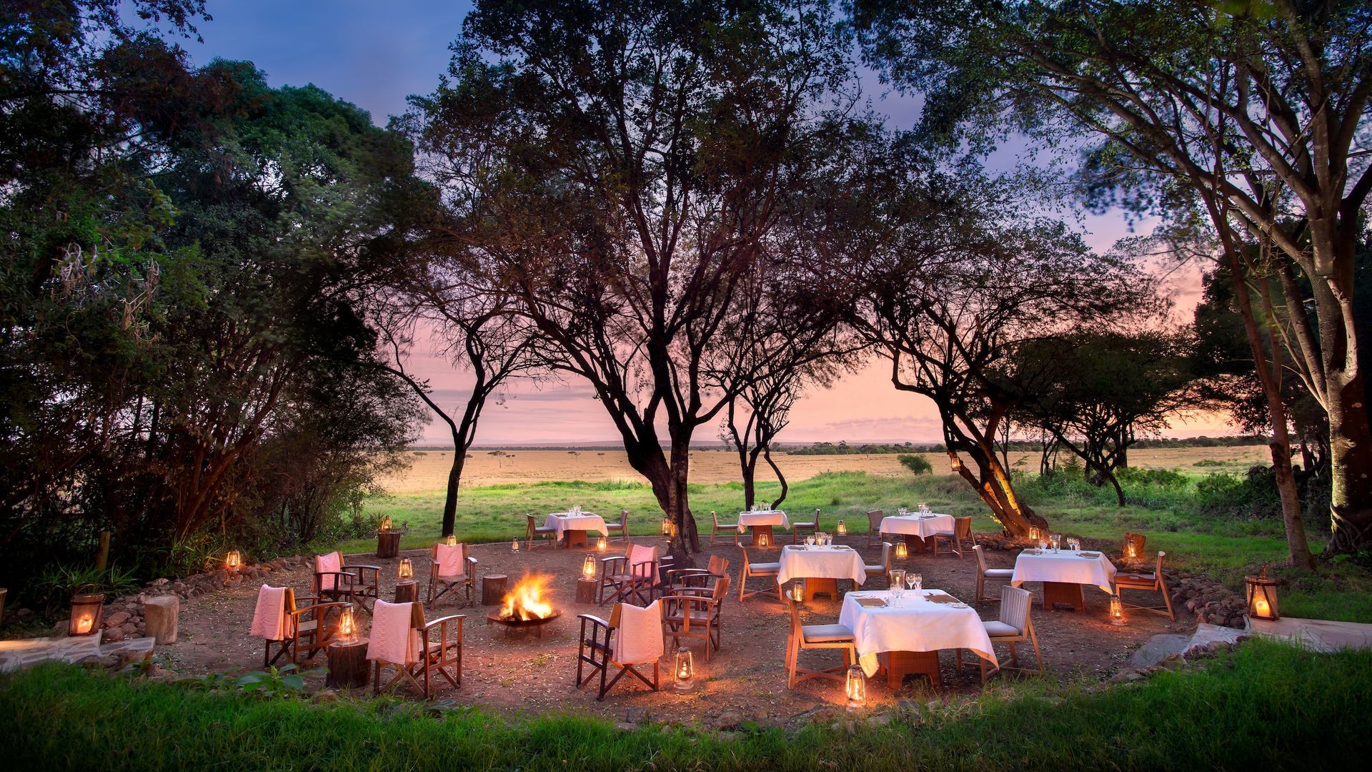 An outdoor dining area at sunset with tables, chairs, and a fire pit surrounded by trees in a savanna setting.