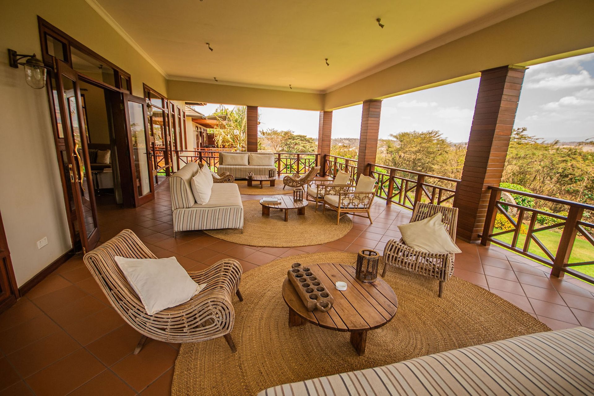 A wide, covered terrace featuring several seating areas with rattan chairs, sofas, and wooden tables on circular rugs.