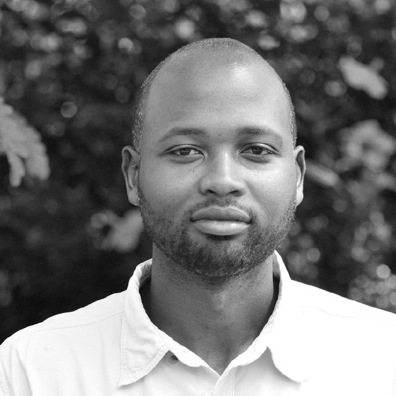 A portrait of a person with a beard, wearing a collared shirt, set against an out-of-focus background of foliage.