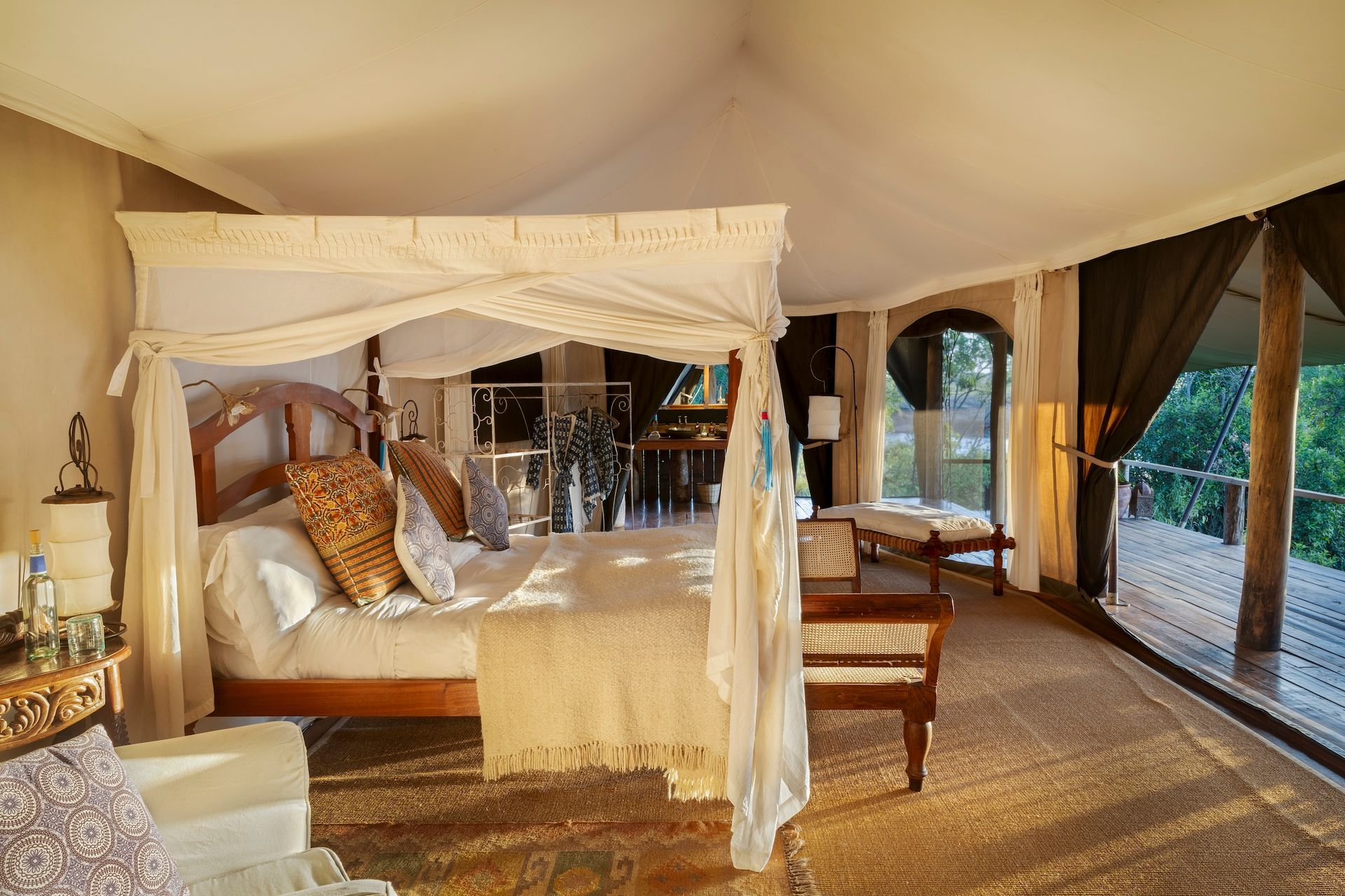 A luxurious safari tent bedroom with a four-poster bed, neutral tones, wood furniture, and a private wooden deck view.