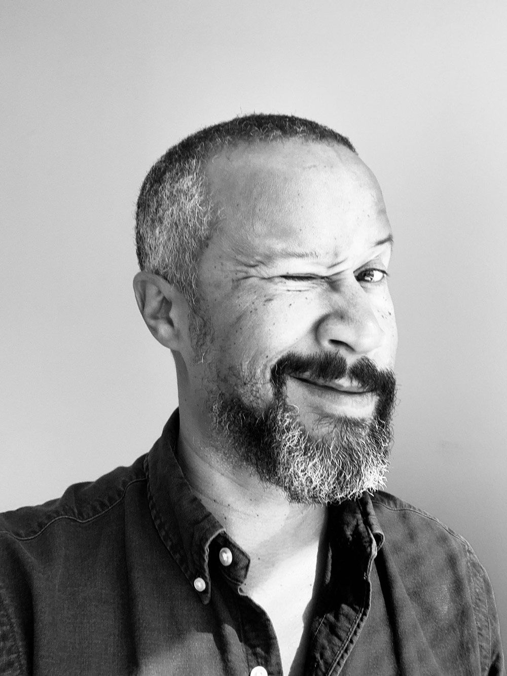 Black and white portrait of a person wearing a button-down shirt, looking at the camera with a playful wink and smirk.
