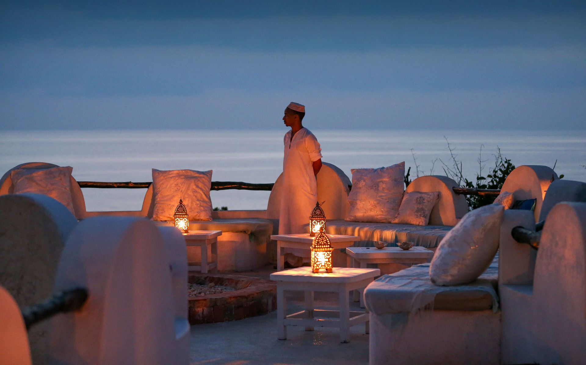 A person stands on a terrace overlooking the ocean at dusk, surrounded by illuminated lanterns and cushioned seating.