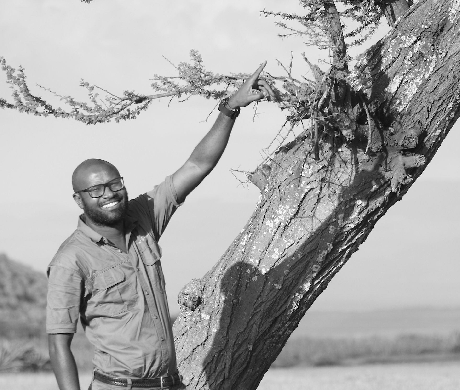 A smiling person in a button-down shirt stands next to a large, leaning tree, pointing toward its branches.