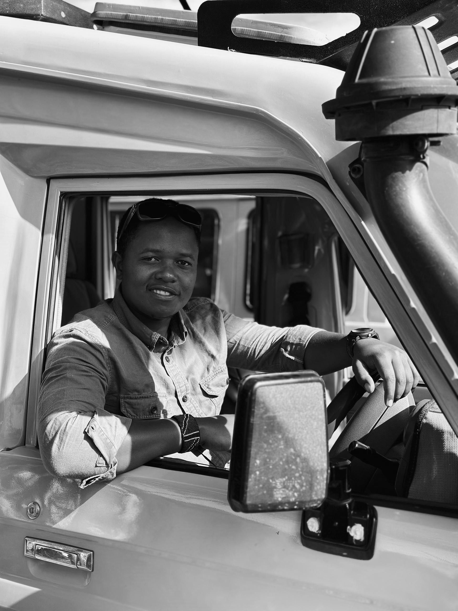 A person sits in the driver’s seat of a vehicle with a roof rack and snorkel, smiling at the camera.