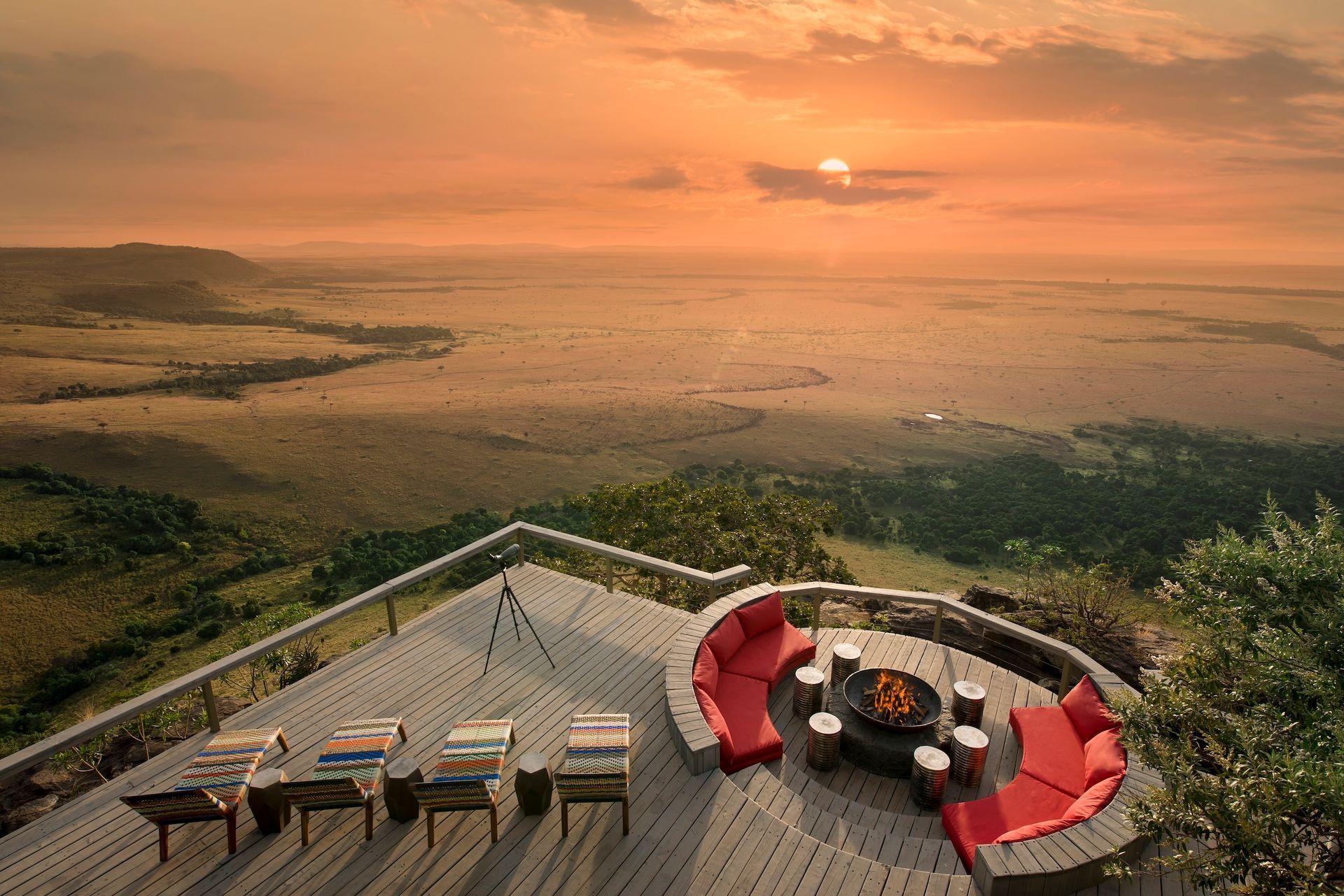 A sunset view from an elevated wooden deck overlooking an expansive African savanna with red seating and a fire pit.