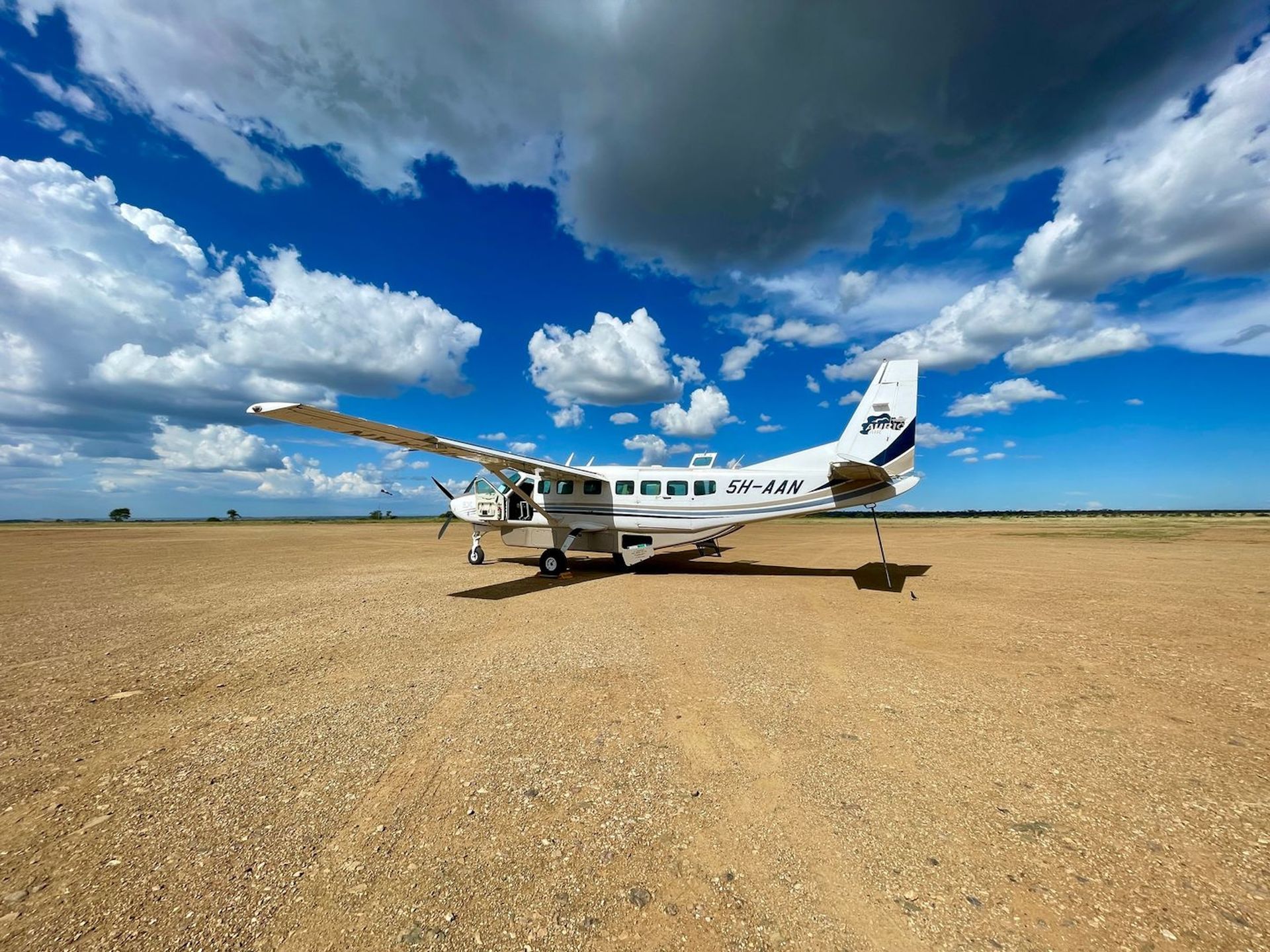 A white bush plane parked on a vast, dry, rocky desert landscape under a blue sky with fluffy clouds.