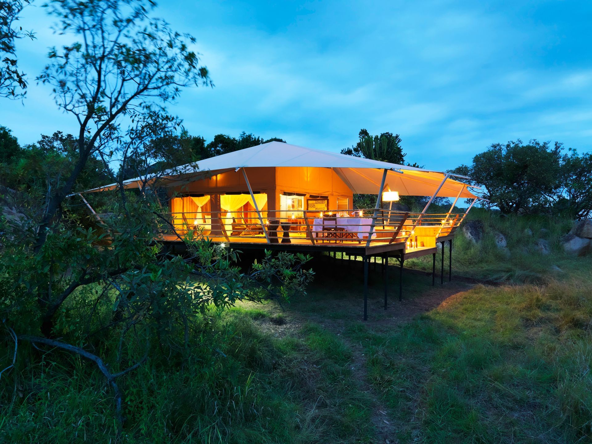 An illuminated safari tent structure on an elevated wooden deck at dusk, surrounded by trees and grassy plains.