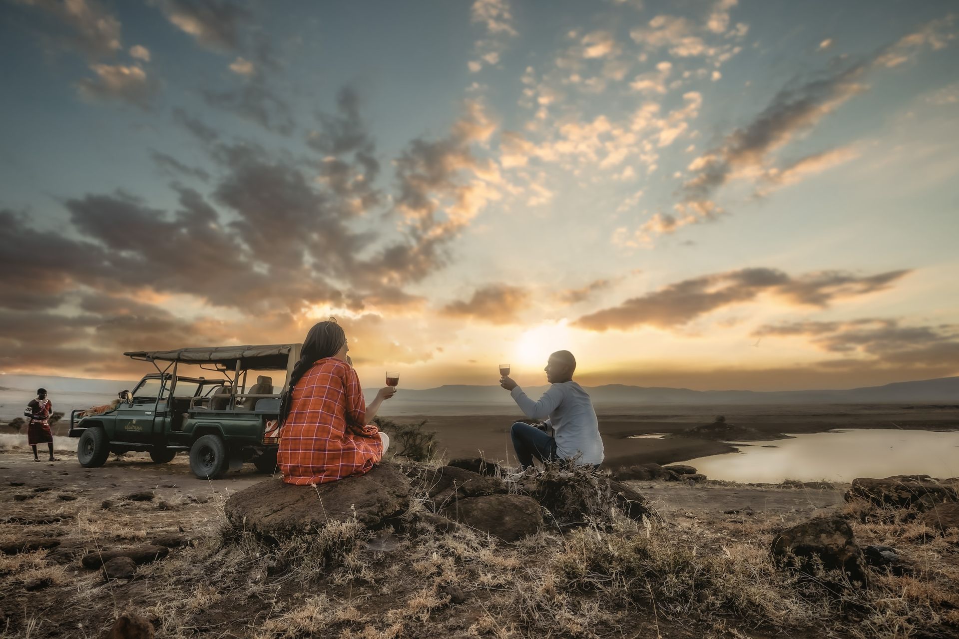 Two people sit on rocky ground in a safari landscape, holding glasses while toasting toward the sunset near a jeep.