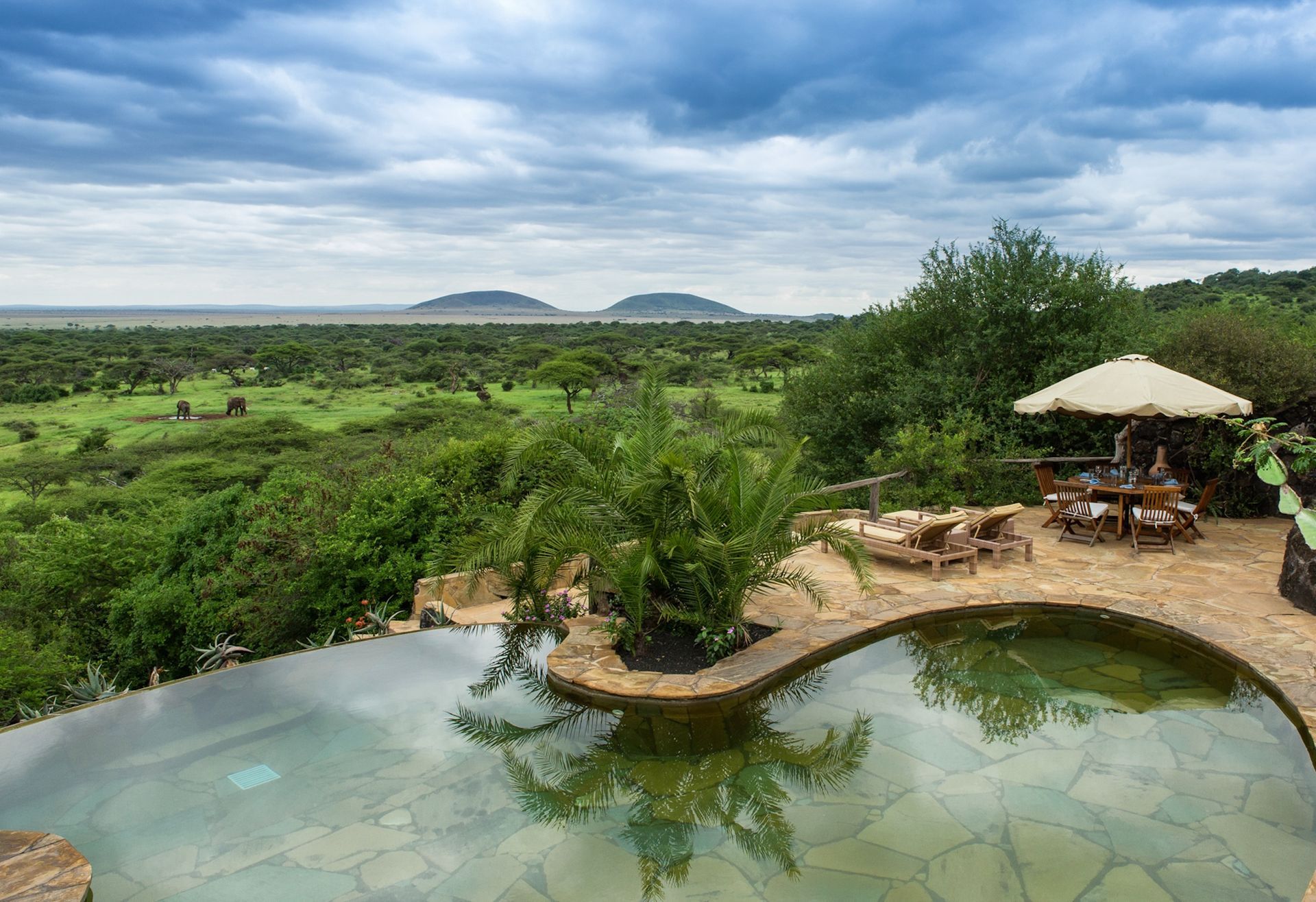 An infinity pool overlooking a vast, green savanna with distant hills under a cloudy sky, featuring patio seating nearby.