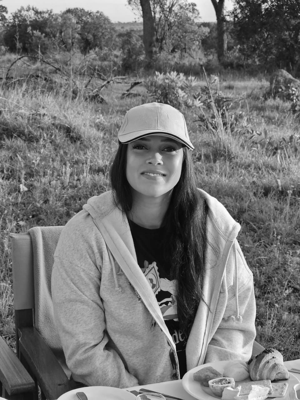 A person in a baseball cap and hoodie sitting at a table with food outdoors, smiling at the camera.