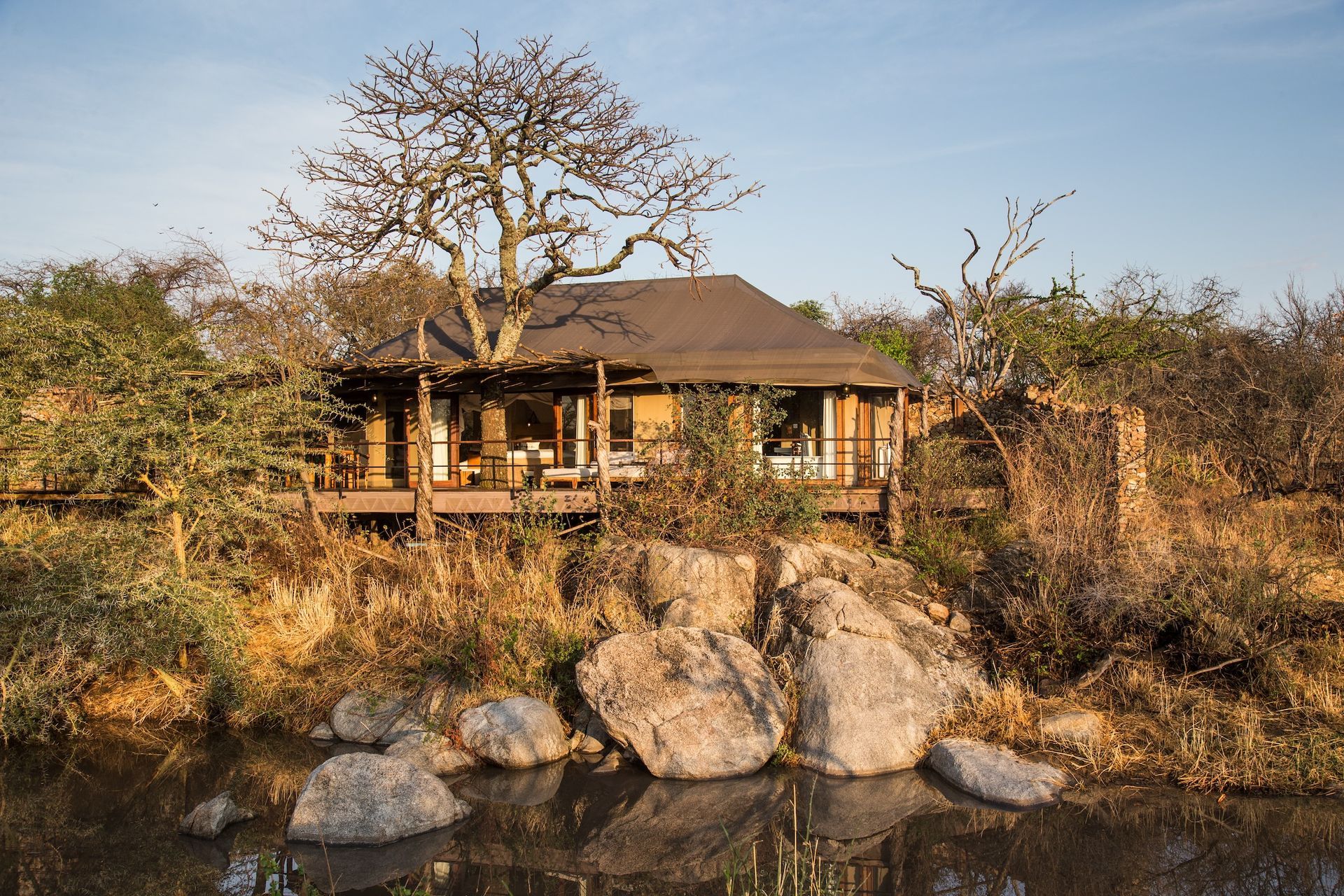 A safari lodge with a dark roof and wooden deck stands on a rocky riverbank beneath a large tree in a dry landscape.