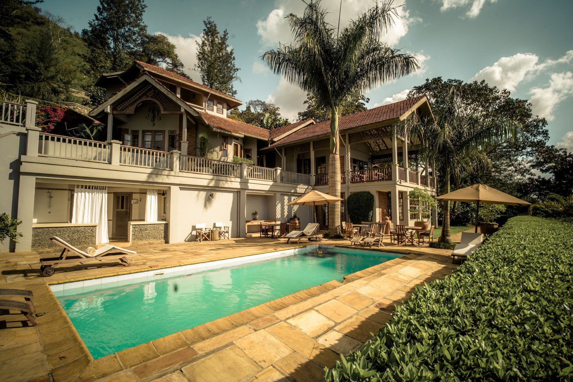 A sunny, two-story villa with a tiled roof features a swimming pool on a stone patio, surrounded by trees and greenery.