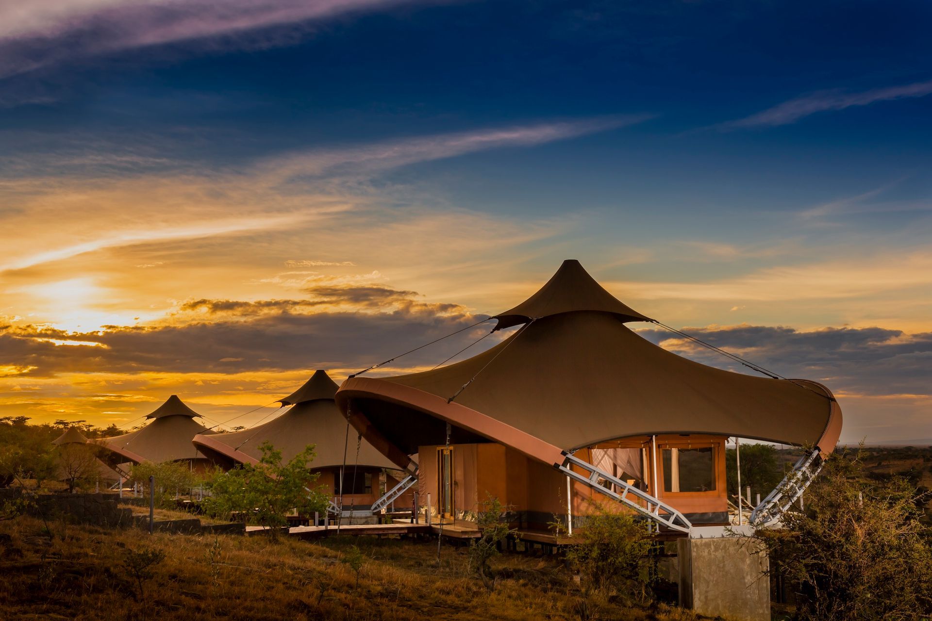 Luxury safari tents with tan canvas roofs set on a hillside against a dramatic sunset sky.