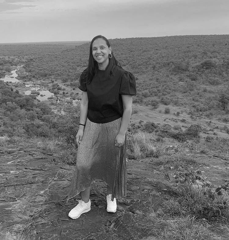 A person standing on a rocky overlook, smiling, with a lush, hilly landscape and a river in the background.