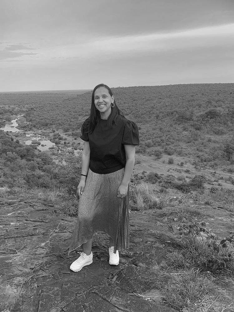 A smiling person in a dark top and patterned skirt standing on a rocky hill overlooking a vast, wooded landscape.