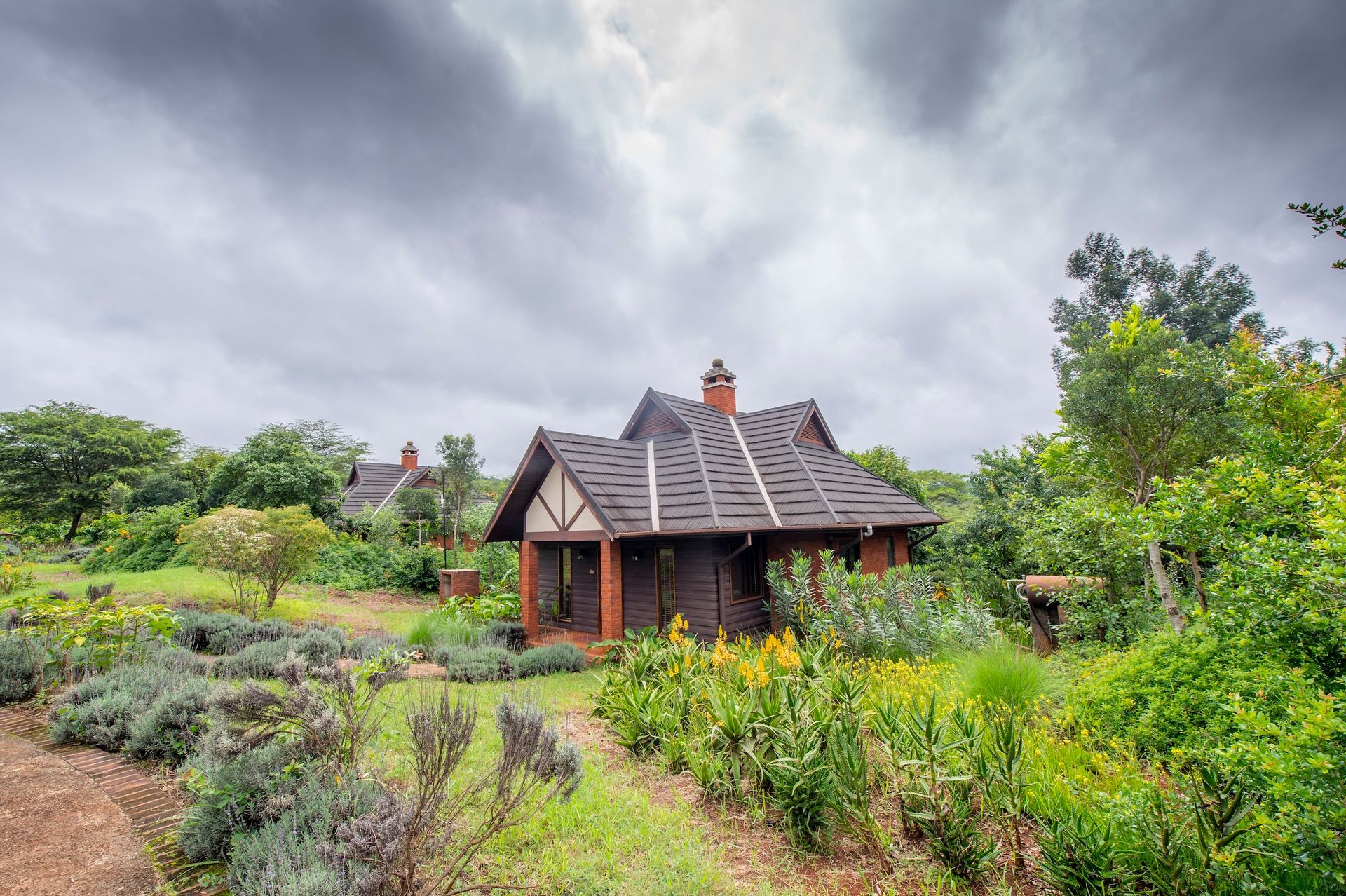 A rustic, dark-wood cabin with a steep roof sits in a lush garden under a dramatic, cloudy sky.