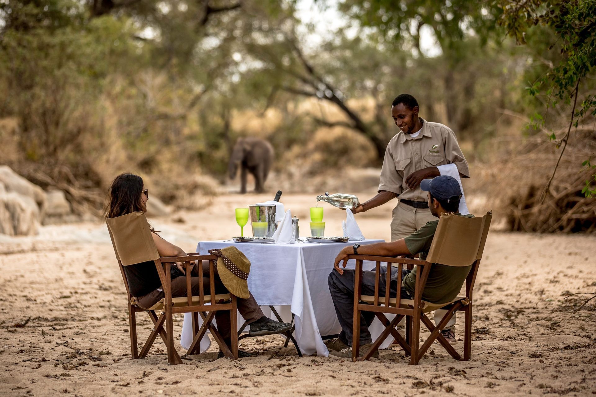 A server pours a drink for two people having an outdoor meal on a sandy riverbank, with an elephant standing in the back.
