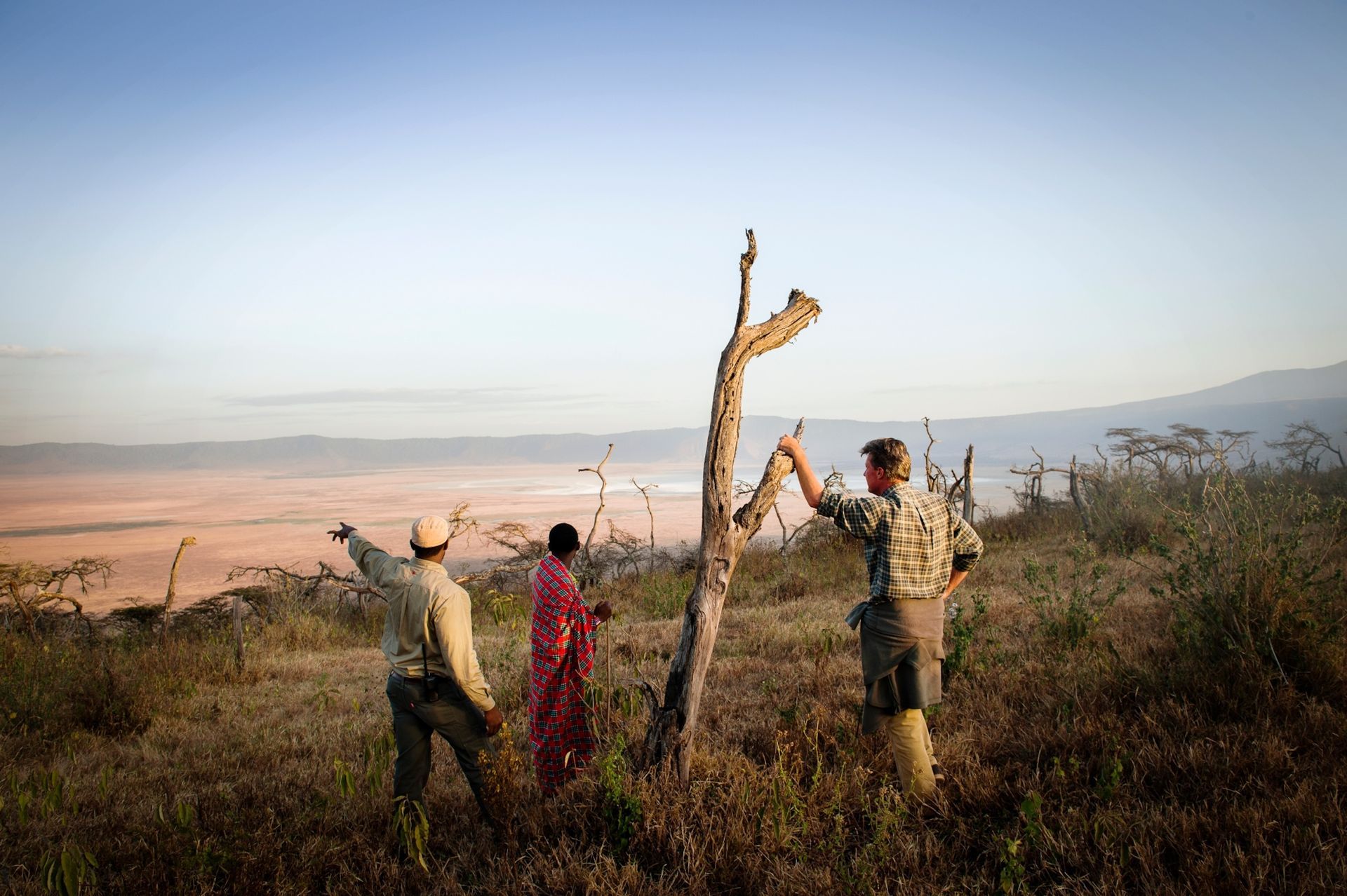Three people stand on a grassy ridge overlooking a vast savanna, pointing toward the landscape at sunset.