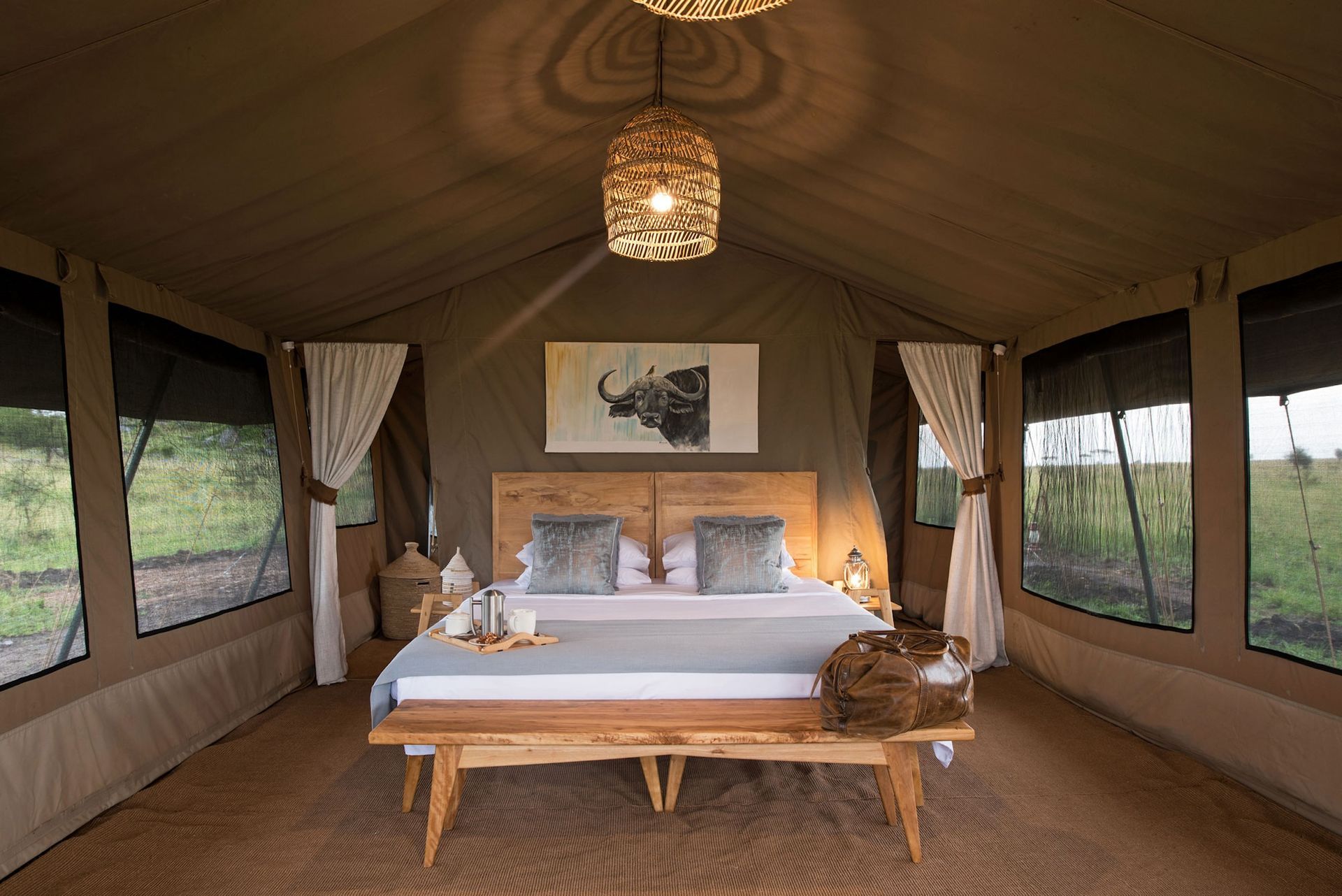 A luxury safari tent interior with a centered king bed, wooden bench, woven pendant light, and framed wildlife art.
