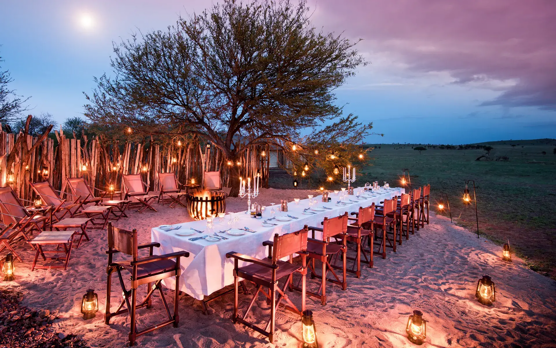 An outdoor evening dining setup on sandy ground under a tree, illuminated by hanging lights and lanterns at dusk.