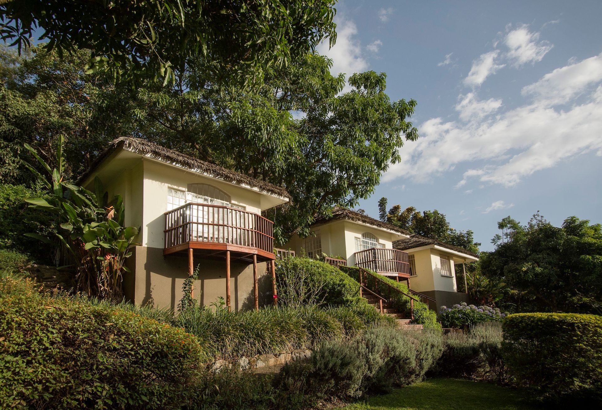 Tropical bungalows with balconies nestled on a lush, green hillside under a blue, partly cloudy sky.