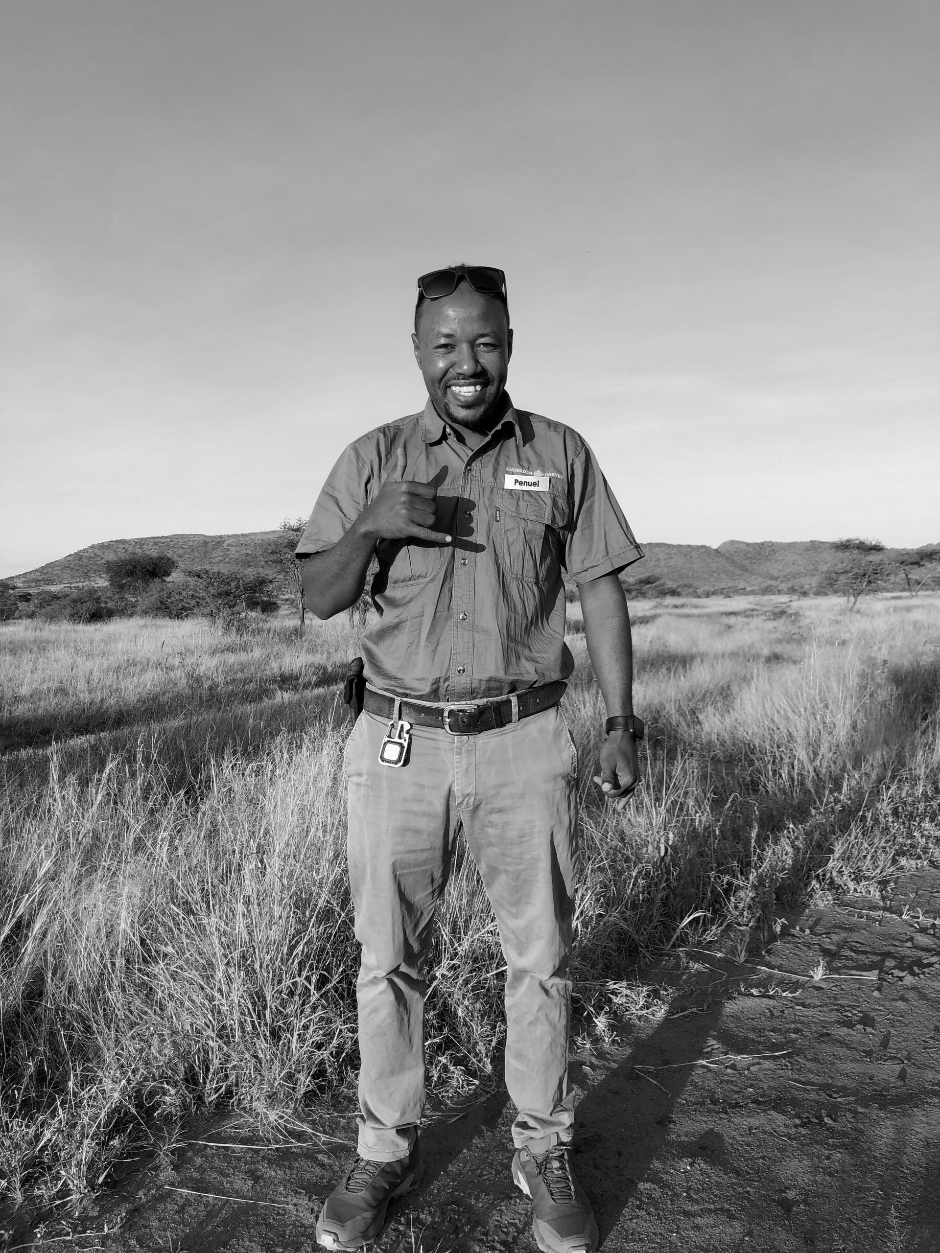 A smiling person stands in a grassy field, gesturing with one hand, wearing a button-down shirt and pants.