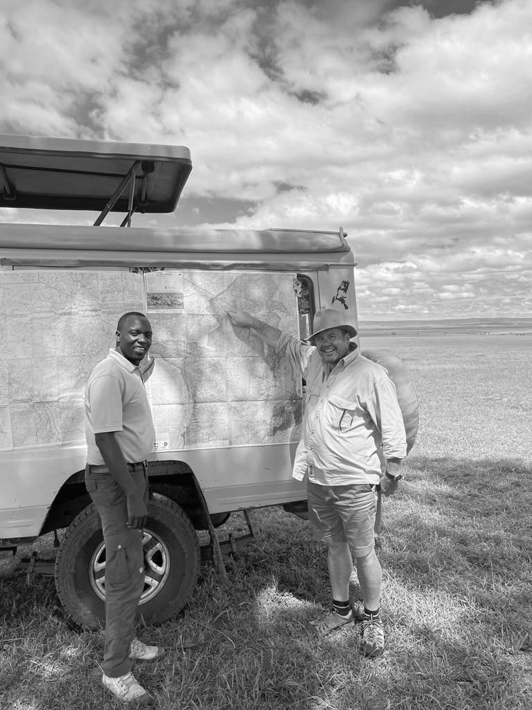 Two people stand next to a safari vehicle in an open, grassy landscape under a cloudy sky.
