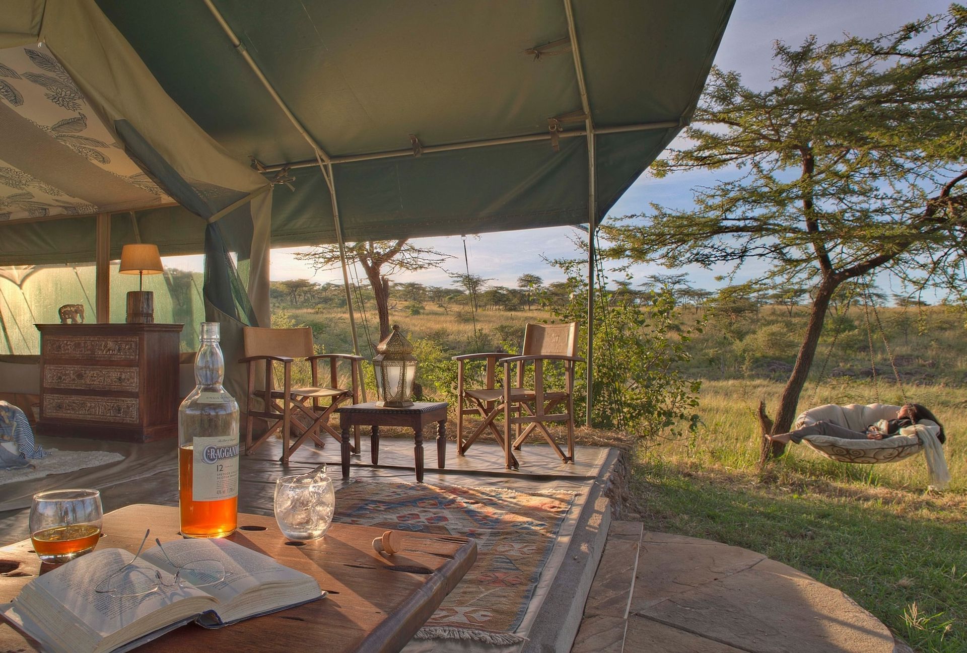 A tented safari deck overlooks a grassy savanna with a bottle of whiskey, a book, and chairs at sunset.
