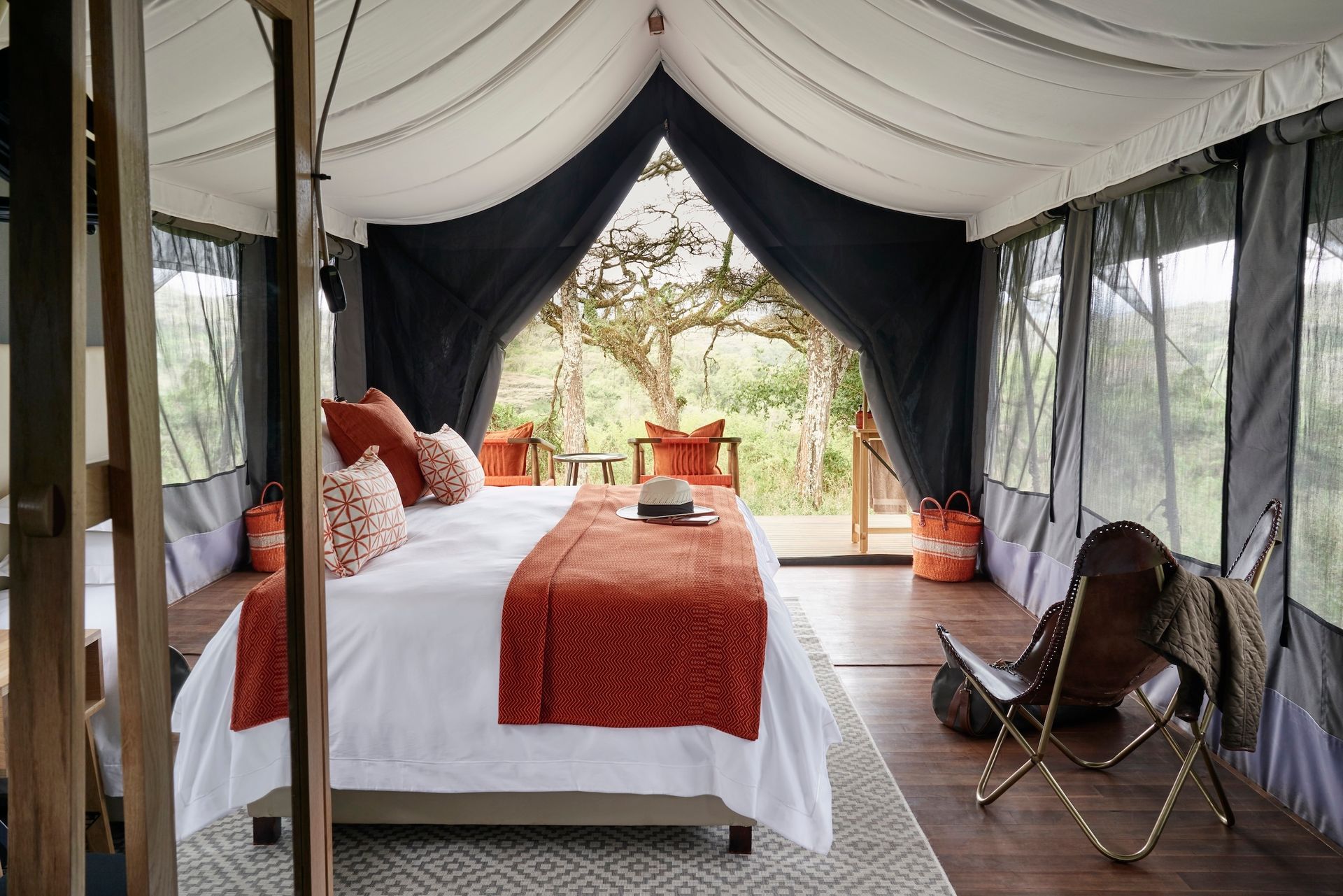 A luxury safari tent interior with a bed featuring orange accents and a folding leather chair, opening onto a wooded view.
