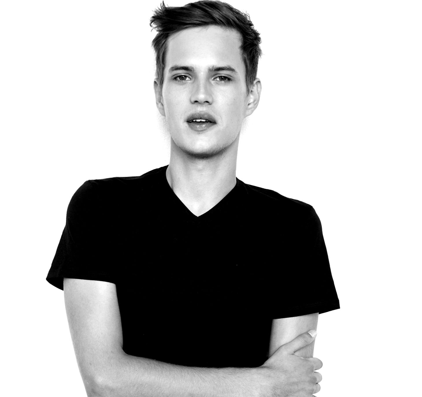 A person with short, dark hair wearing a black V-neck shirt, posing against a white background with arms crossed.