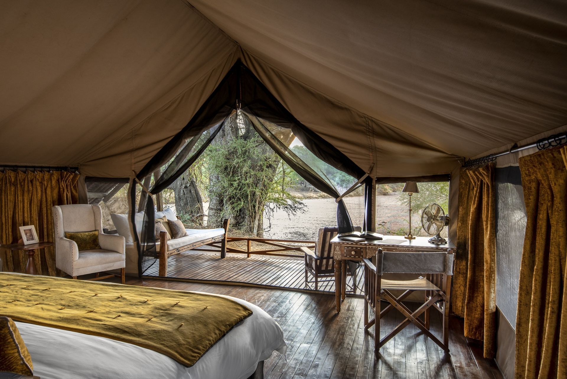 A luxury safari tent interior with a bed, white chair, and desk looking out onto trees through an open mesh screen.