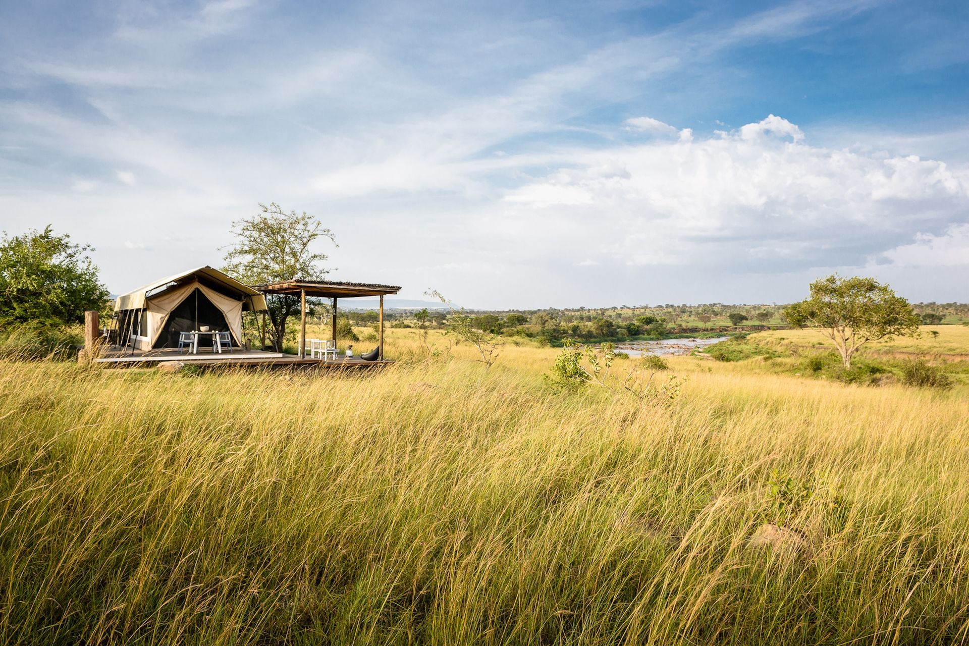 A canvas tent structure on a wooden deck overlooks a vast, golden savanna under a bright, partly cloudy sky.