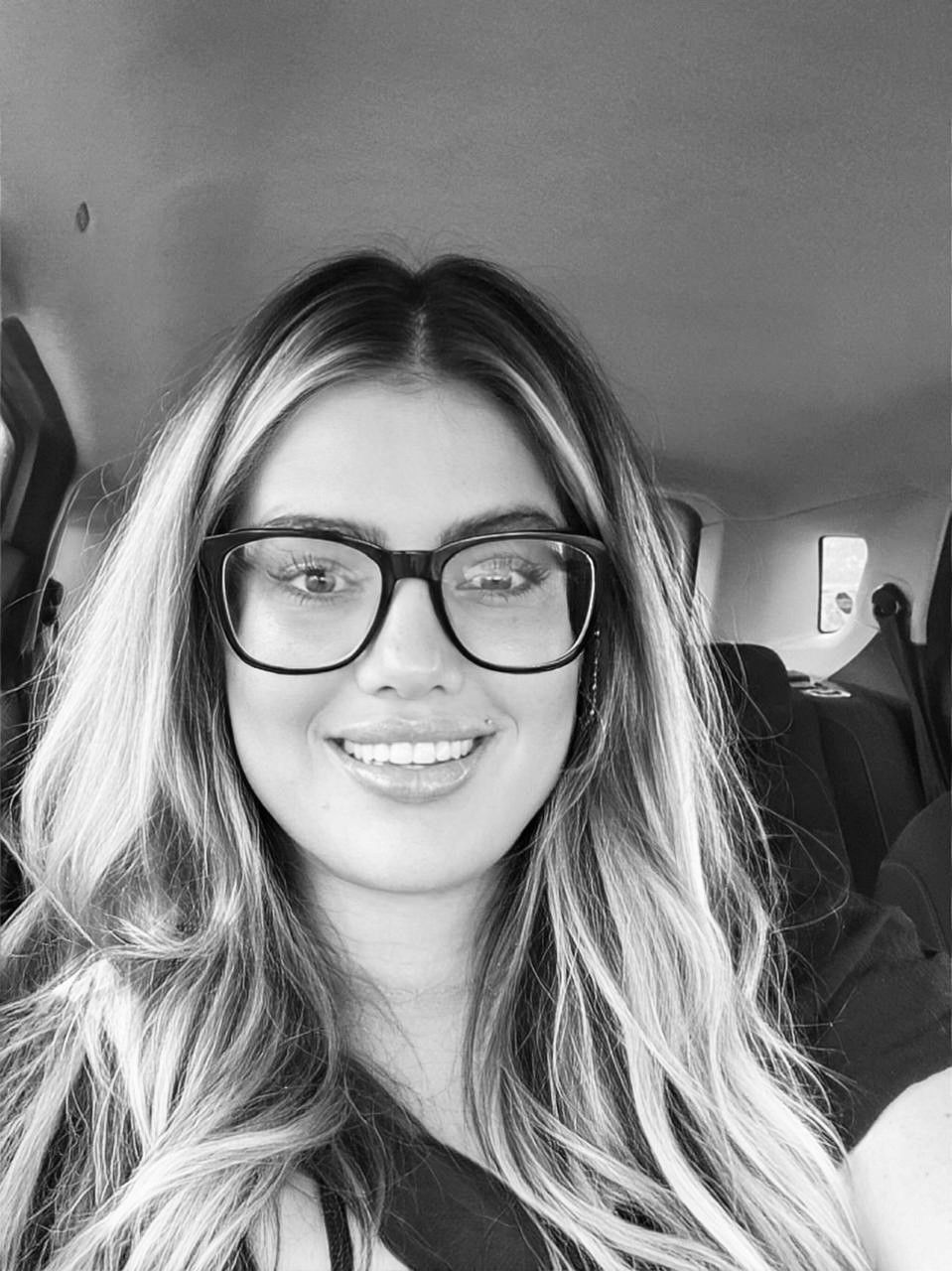 A smiling person with shoulder-length, highlighted hair wearing thick-rimmed glasses in the backseat of a car.