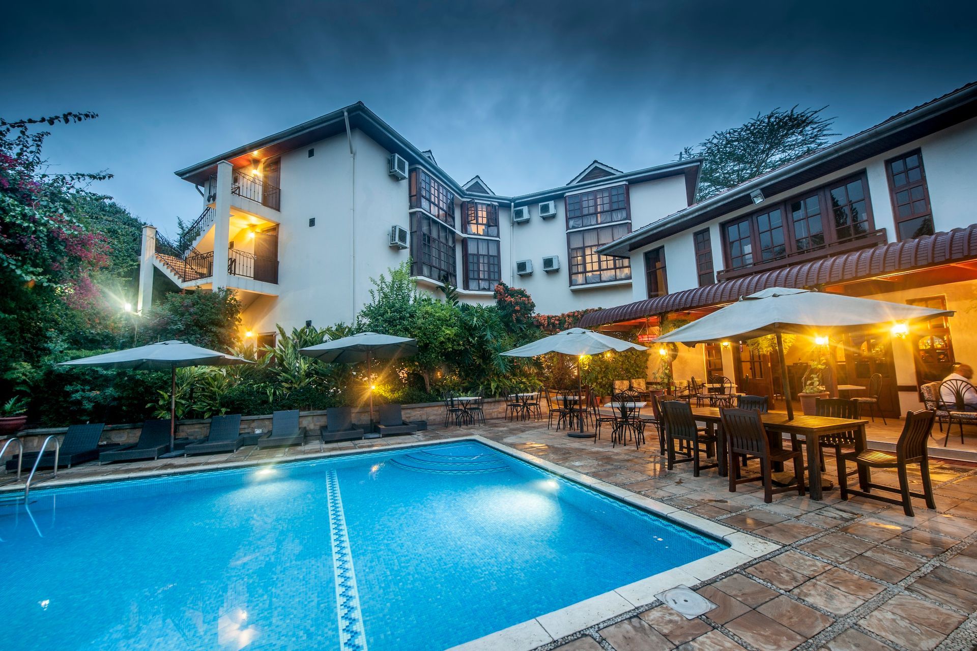 A swimming pool at a resort at twilight, with outdoor dining tables under umbrellas and a multi-story building.