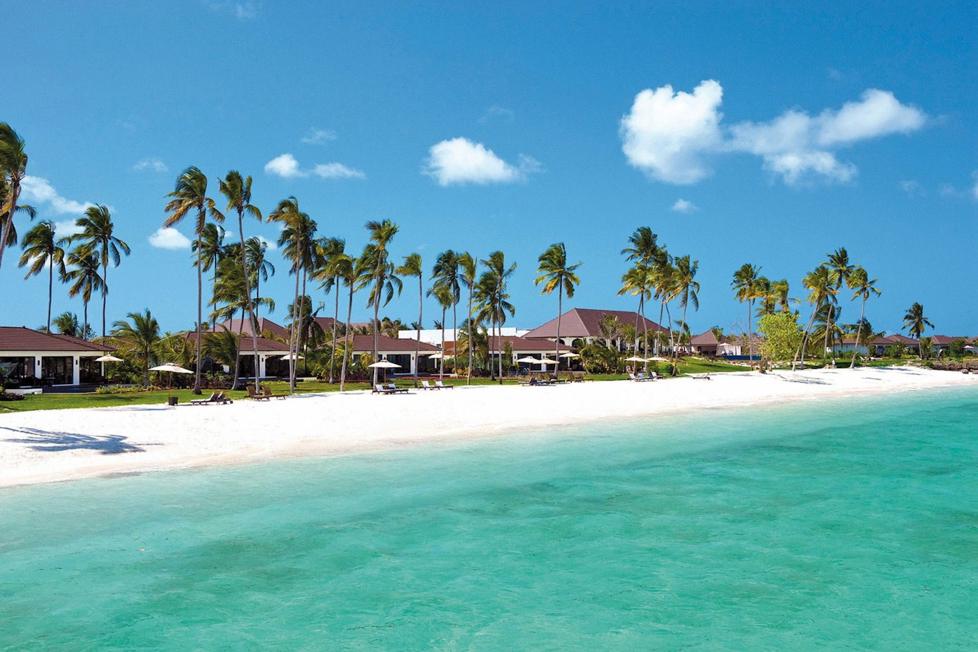 A white sand beach with turquoise water, palm trees, and tropical resort buildings under a clear blue sky.