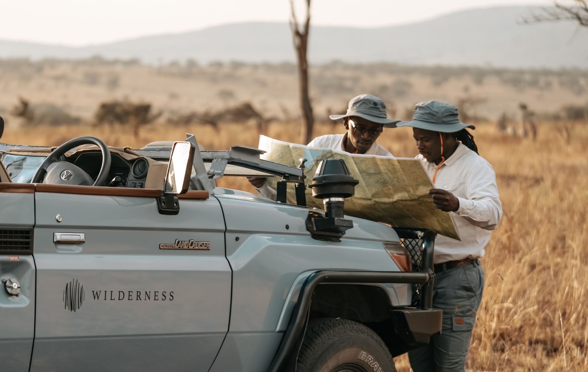 Two guides in hats study a map over the hood of a blue safari vehicle in a dry, grassy savanna.