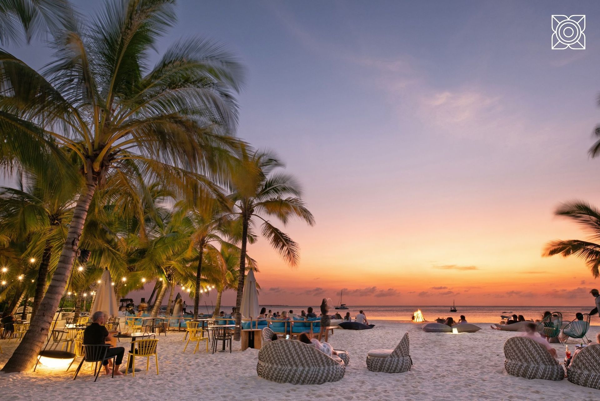 People relax on a white sand beach at sunset, with palm trees, outdoor lights, and wicker lounge chairs under a soft sky.
