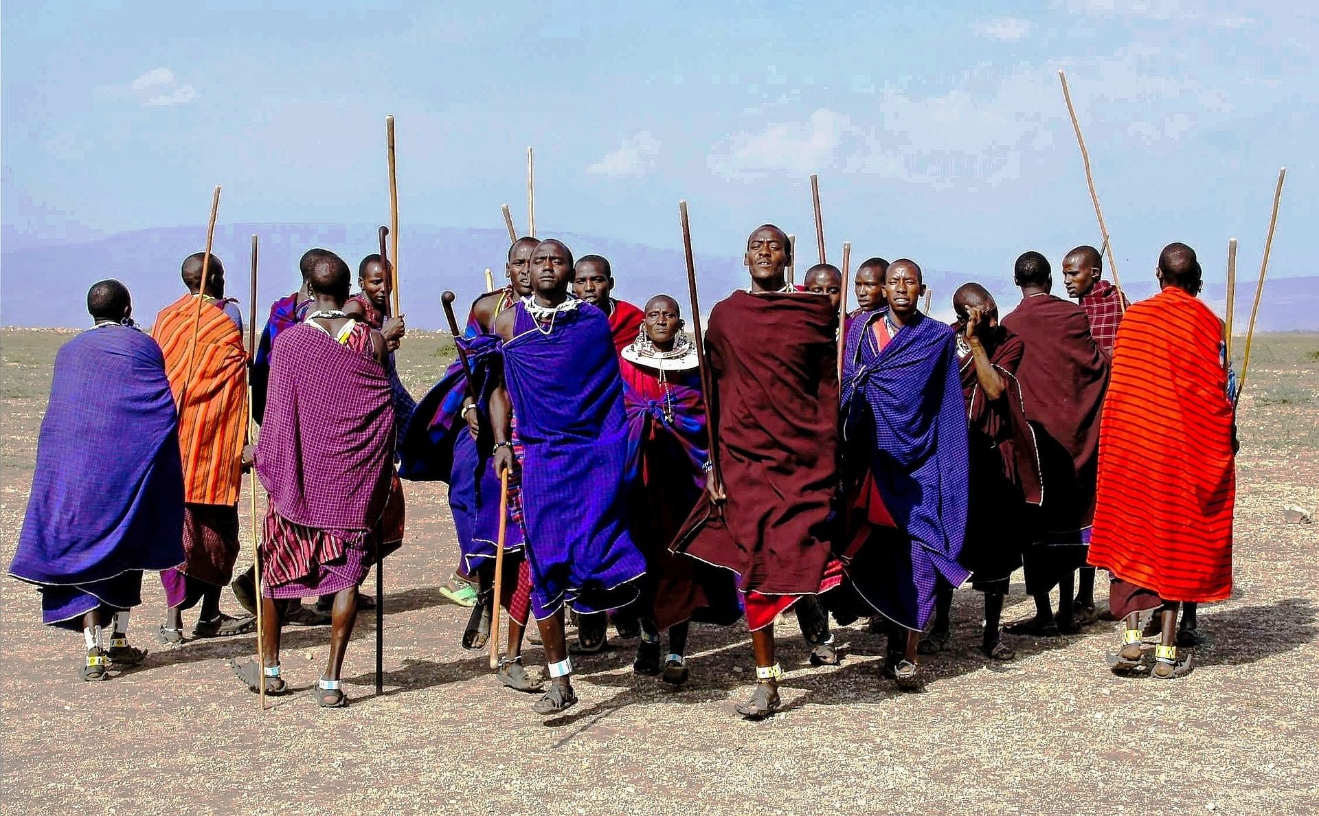 A group of people wearing traditional red, blue, and purple shuka cloths stand in an open, arid landscape holding staves.