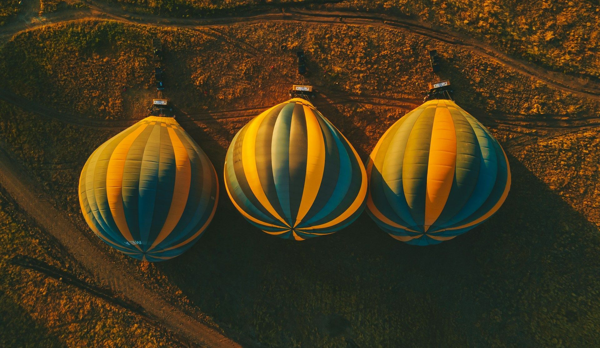 An aerial view of three striped yellow and blue hot air balloons resting on a dark, textured field at sunrise.