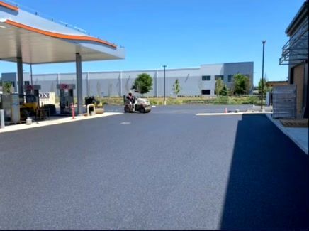 Black asphalt being rolled at a gas station with a large building in the background on a sunny day.