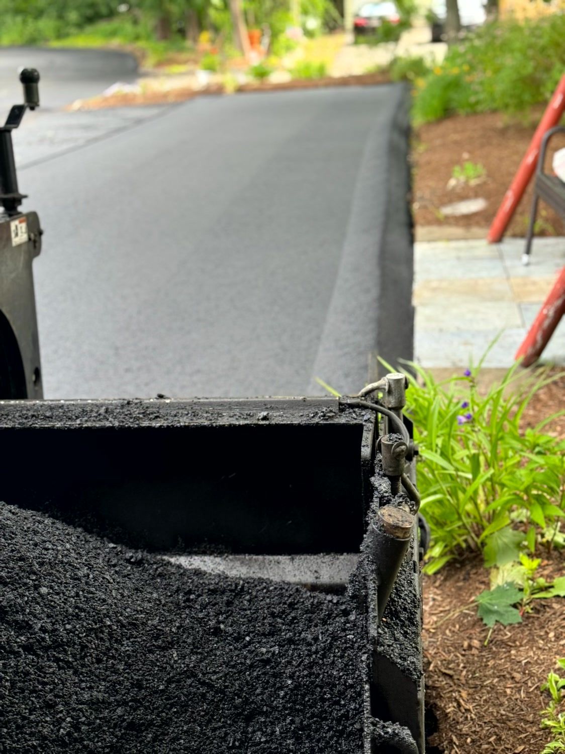 Freshly paved asphalt road alongside a garden bed; a truck bed filled with asphalt in foreground.