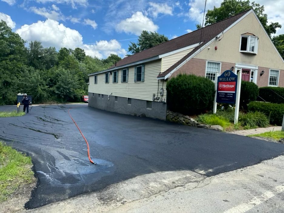 Asphalt parking lot being sealed next to a building on a sunny day. A worker is seen applying the seal.