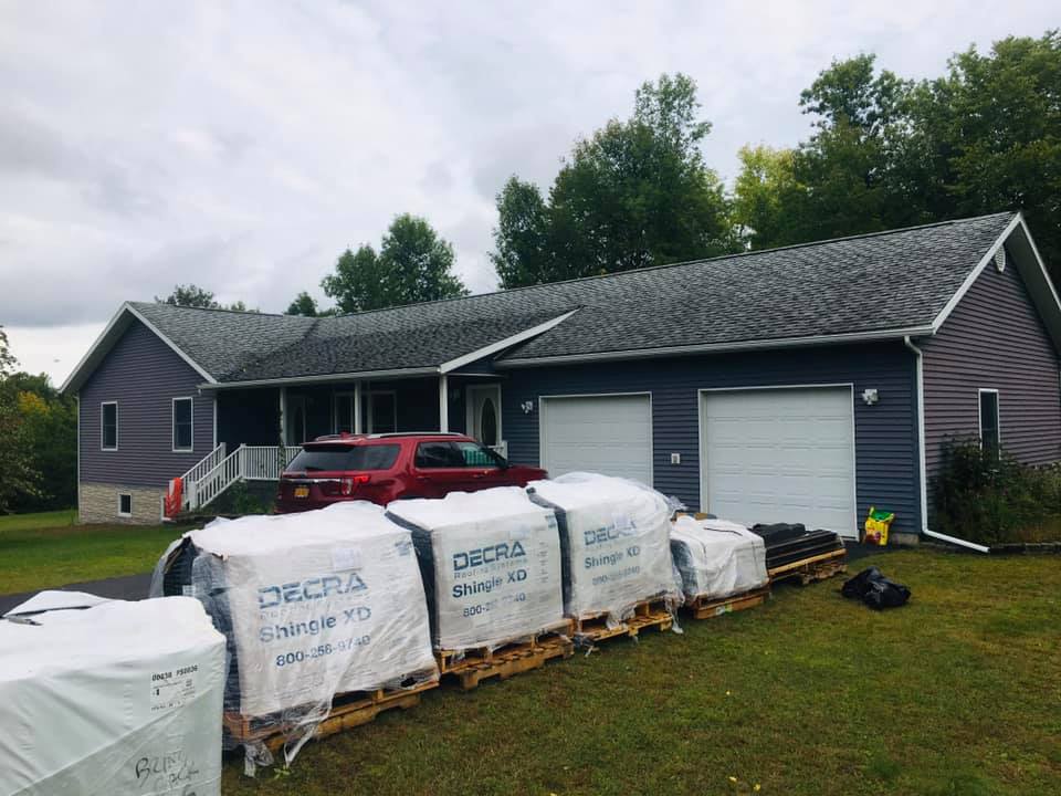 House with roof under repair, pallets of roofing materials in the front yard, and a red truck parked by the porch