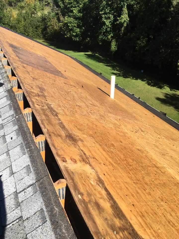 Wooden roof sheathing under construction beside gray shingles, with a white vent pipe visible.