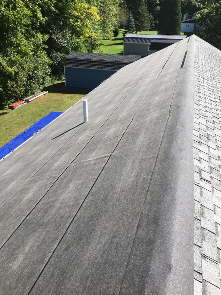Long gray rooftop with skylights beside green trees and blue tarps