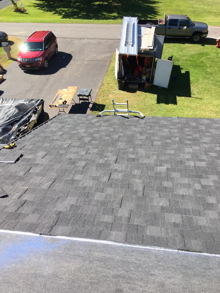 Aerial view of a shingled roof with vehicles, a dumpster, and roofing materials on a lawn below.